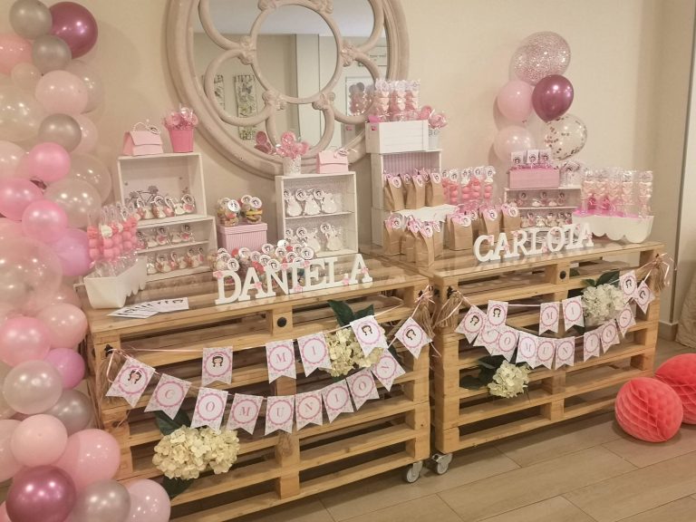 Mesa dulce de comunión montada sobre palés, con globos rosas y los nombres de las niñas